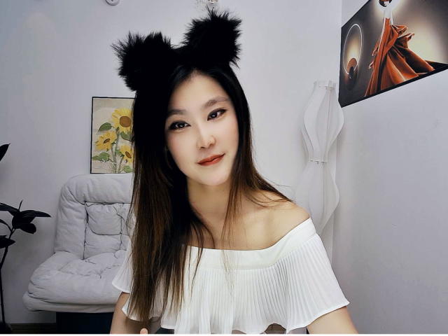 AilsaJade with black furry cat ears, wearing a white off-shoulder top, has long brown hair, red lipstick, and a cute smile, in a white room with a chair.