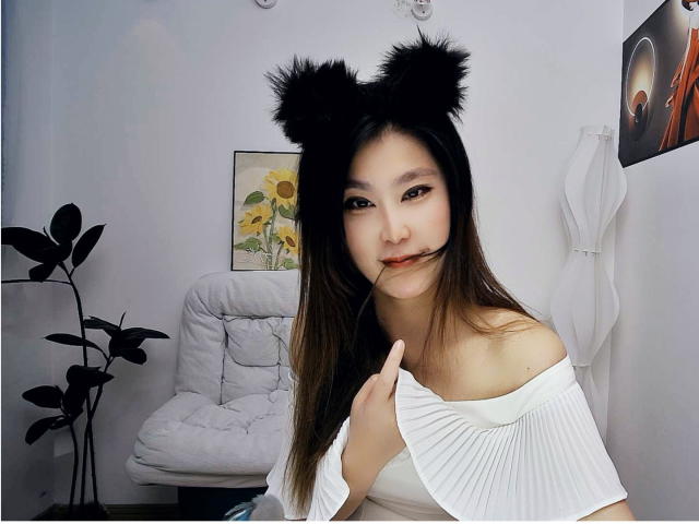 AilsaJade with black furry cat ears, biting her finger, wearing a white off-shoulder top, holding her shoulder, long brown hair, white chair and plant in background.
