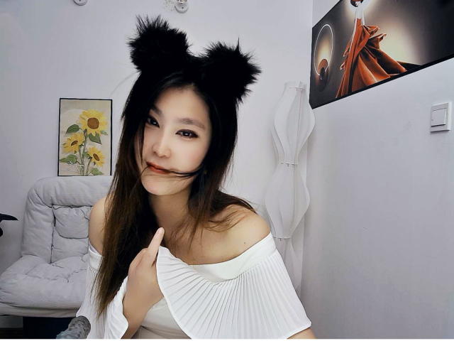 AilsaJade with black furry cat ears, long brown hair, white off-shoulder top, holding pleated fabric near tits, smiling with red lipstick, white chair in background.