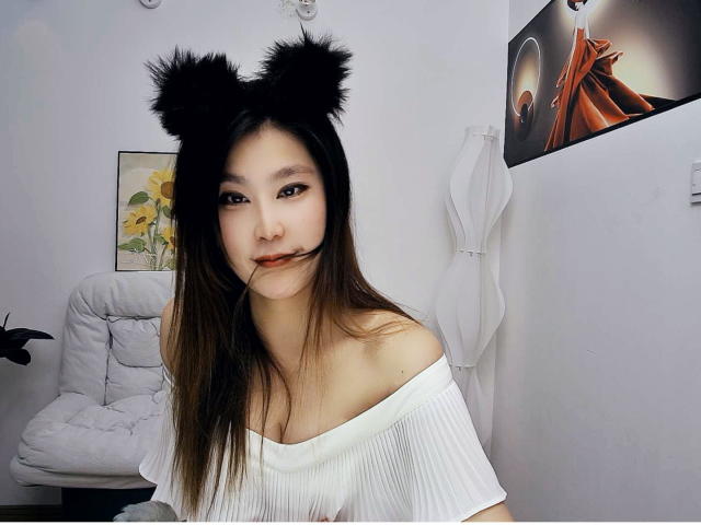 AilsaJade with black furry cat ears, long brown hair, white off-shoulder top, biting her hair, white wall, white chair, white curtain, and wall art with orange and black.