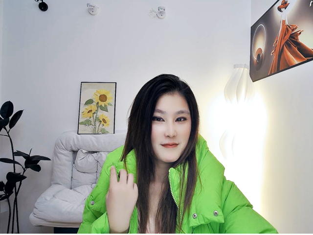AilsaJade with long brown hair, wearing a bright green puffy jacket, has pale skin and a cute face, sitting in front of a white wall with a lamp.