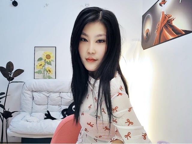 AilsaJade with long black hair, wearing a white dress with orange patterns, sitting on a pink chair, looking at the camera with a slight smile, pale skin, and red lipstick.