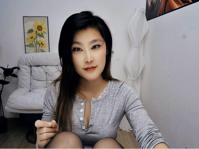 AilsaJade with black hair, light skin, and brown eyes, wearing a grey button-up top, sitting with knees up, looking at camera, in a white-walled room.