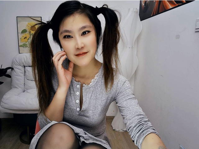 AilsaJade with pigtails, wearing a gray long-sleeve shirt and black tights, holding a white headset, smiling at the camera, sitting on a chair.