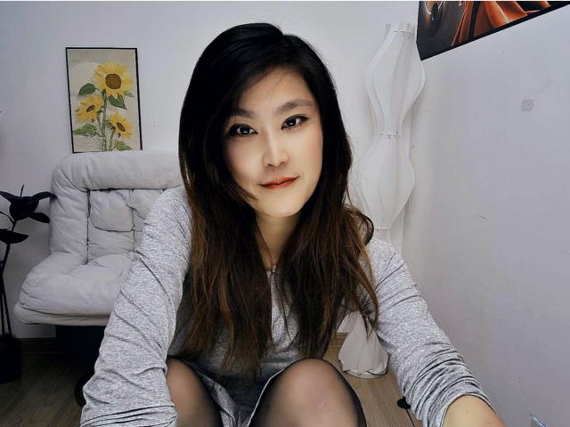 AilsaJade, with long brown hair and light skin, sits on a white couch, wearing a gray long-sleeve top and black shorts, smiling at the camera. Her pussy is not visible.