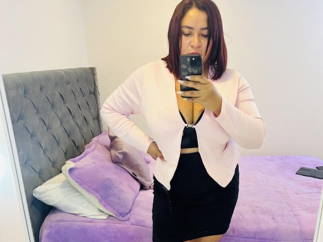 KarlaWild with red hair, pink cardigan, black bra, and black skirt, standing in front of mirror, holding phone, showing her tits, in bedroom with purple bed.