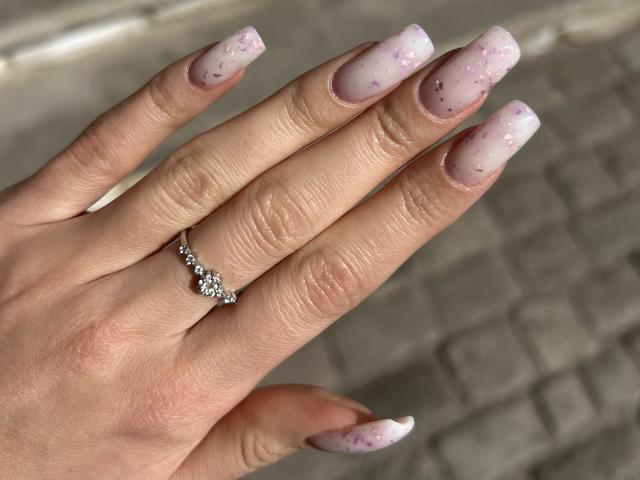 La mano di Karish, con la pelle chiara, lunghi unghie rosa e bianche con brillantini, indossa un anello d'argento con un diamante sul dito anulare..