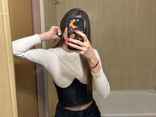 Karish with long brown hair, wearing a white long-sleeve top and black leather corset, holding a sunflower phone case, mirror selfie in a bathroom. Boobs covered by corset.