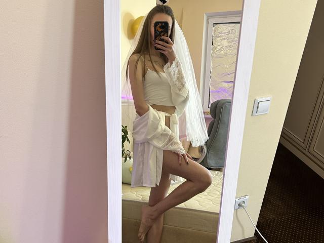 Karish with long blonde hair, wearing a white crop top, sheer white skirt, and a veil, taking a mirror selfie, showing her bare ass and thigh.