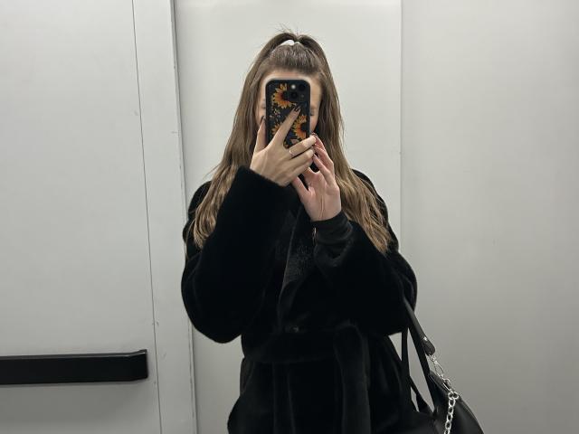 Karish with long brown hair in a ponytail, wearing a black fur coat, holding a phone with a sunflower case, covering her face, standing in a white elevator.