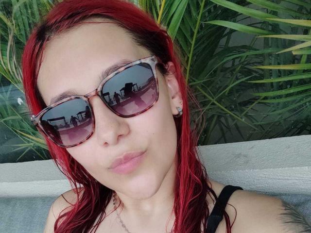 JuliaRouse with red wet hair, wearing purple-tinted sunglasses, black strap top, and small ear studs, has a calm, slightly pouty expression, showing light pink lips.
