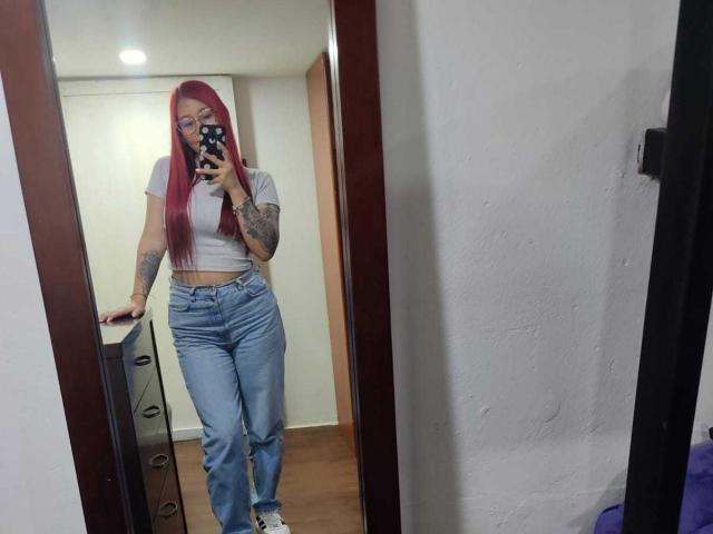 JuliaRouse with red hair, tattoos on her arm, white crop top, blue jeans, standing in mirror selfie, holding phone, white sneakers, wooden floor, white walls, wooden door.