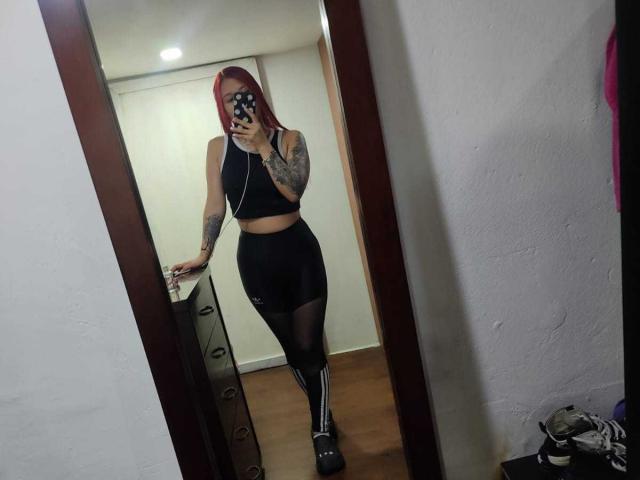 JuliaRouse with red hair, black crop top, black high-waisted leggings, and black knee-high socks, holding phone in mirror selfie, showing tattoos on right arm.