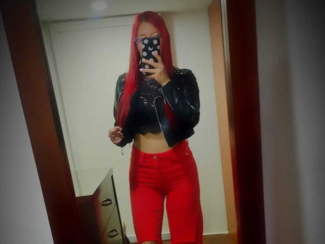 JuliaRouse with long red hair, wearing a black lace top, black leather jacket, and red high-waisted pants, taking a mirror selfie with a polka-dot phone case.