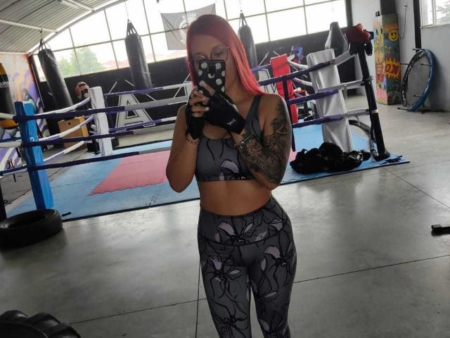 JuliaRouse with pink hair, glasses, black fingerless gloves, gray sports bra, and floral-patterned leggings, taking a mirror selfie in a boxing gym. Her tattooed arm is visible.