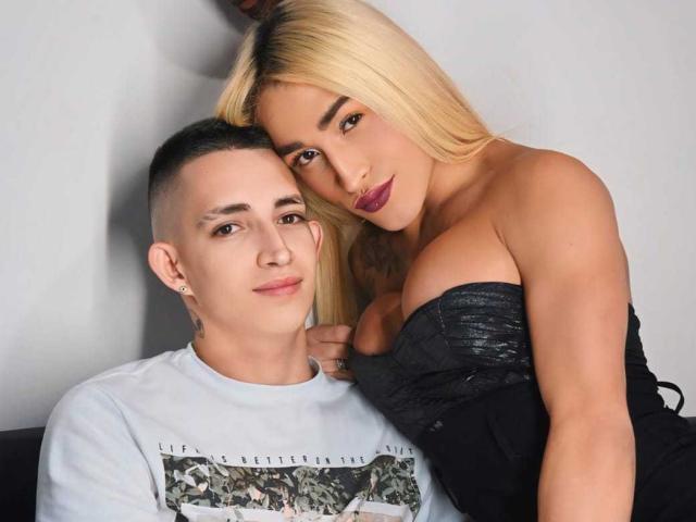 MyaAndJack69 with blonde hair and purple lipstick, leaning on a guy with a shaved head and ear piercings, wearing a black strapless top, showing her tits.