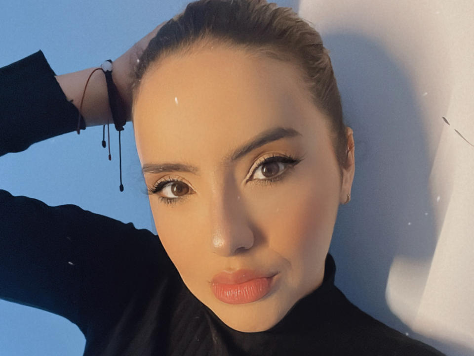 BelleSuavage has light brown skin, brown eyes, and red lipstick. She's wearing a black top, with her hand in her hair, and a black bracelet. She's looking at the camera.
