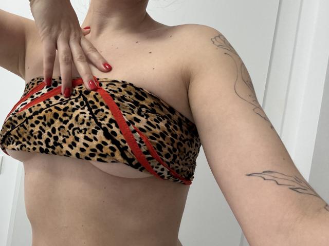 EricaLust's titties are covered by a leopard-print bra with red trim, showing tattoos on her arms, and she's got red nails on her hand.
