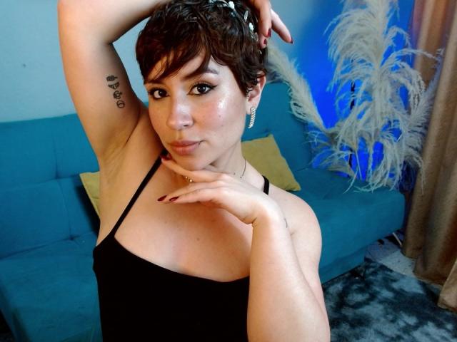 Sara Kleine, com cabelo castanho curto, top preto sem alças, braço levantado mostrando uma pequena tatuagem de coração, mão no queixo, sofá azul, travesseiro dourado, cortinas bege e penas brancas fofas..