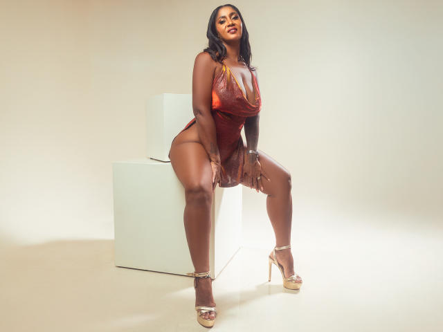 CristinaWillis is a dark-skinned woman with large tits, wearing a red, sheer, crotchless dress, sitting on a white cube, in gold high heels, with a sexy smile.