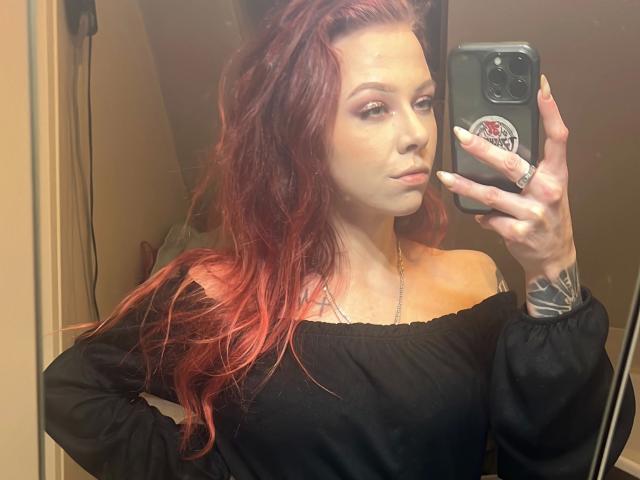 MeriJey-hot with red hair, off-shoulder black top, holding phone in mirror selfie, showing tattoo on wrist, subtle makeup, confident expression, slim build, sexy, reflection visible.
