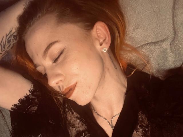 MeriJey, con los ojos cerrados y vistiendo una blusa negra de encaje, tiene el cabello naranja, un tatuaje en el brazo y pequeños pendientes de diamantes, está acostada con la mano sobre la cabeza..