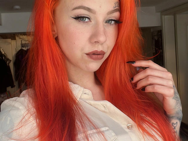 MeriJey-hot with bright orange hair, black eyeliner, and pink lips, showing off black nails, tattoo on her right hand, and wearing a white button-up shirt.