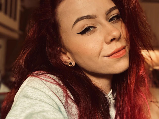 MeriJey-hot a les cheveux roux, un trait d'eye-liner noir, du rouge à lèvres orange et un léger sourire. Elle porte une chemise vert clair et des boucles d'oreilles dorées. Sa peau est claire..