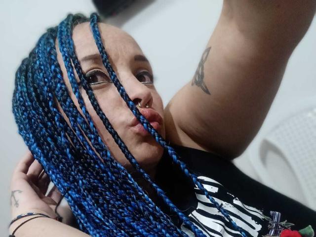 Dalila Musa mit blauen Zöpfen, voller Lippen, Nasenring, Brille, schwarzem T-Shirt, Tattoo am Arm, Selfie, weißer Plastikstuhl im Hintergrund, hält sich das Haar, blickt in die Kamera..