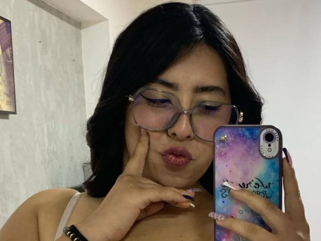 ScarlettParchMusa with dark hair, glasses, and purple lipstick, pouting, holding a phone with a galaxy design, touching her cheek, showing her tits, and wearing a white bra.