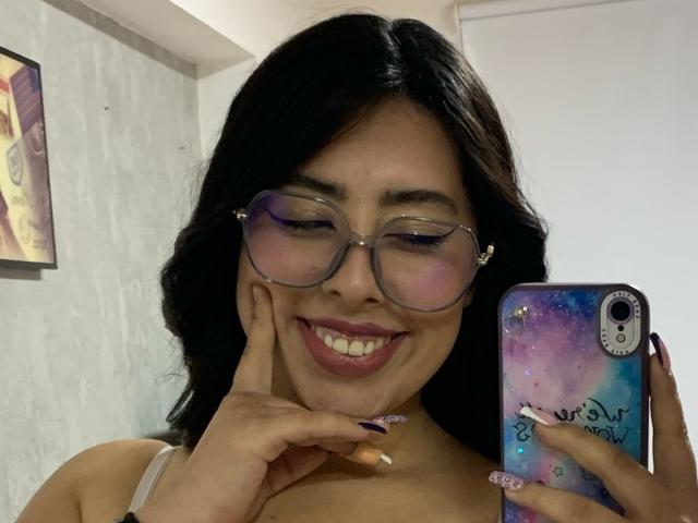 ScarlettParchMusa with dark hair, glasses, smiling, holding phone with galaxy case, fingers on cheek, showing off pink and purple nails, white bra, cute, Asian, attractive.