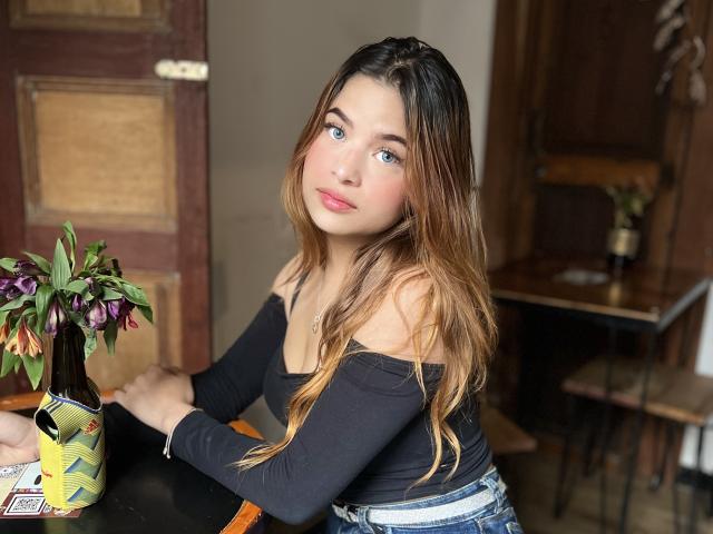 ShantalSummer with blue eyes, long brown hair, wearing a black off-shoulder top and blue jeans, sitting at a table with a beer bottle and flower in a vase.