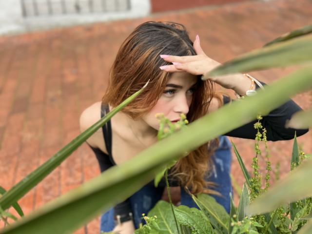 Shantal, de cabello castaño, con jeans azules y una blusa negra, arrodillada, con una mano en la cabeza, mirando hacia un lado, una planta verde en primer plano, suelo de ladrillo rojo, fondo borroso..