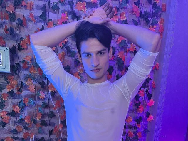 DavidKarr with arms raised, wearing a sheer white shirt, revealing small nipples, in a purple-lit room with leaf-patterned wallpaper. He has dark hair and a serious expression.