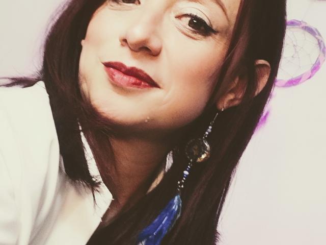CatalinaMonyque's face, slight tilt, brown hair, red lipstick, small nose, blue earrings with purple and blue beads, white shirt, soft smile, light skin, subtle makeup, gentle expression.