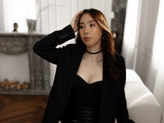 AnnaReyla with brown hair, black choker, black strapless top, black blazer, hand in hair, small tits, tattoos on chest, sitting on white couch, wooden floor, relaxed expression.