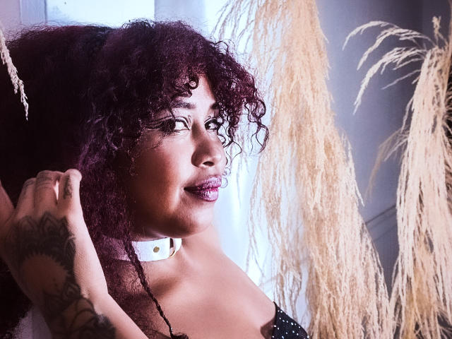 DalilaHeinz with curly purple hair, dark skin, tattooed arm, and purple lipstick, looking to the side, black dress with white polka dots, pampas grass in the foreground.