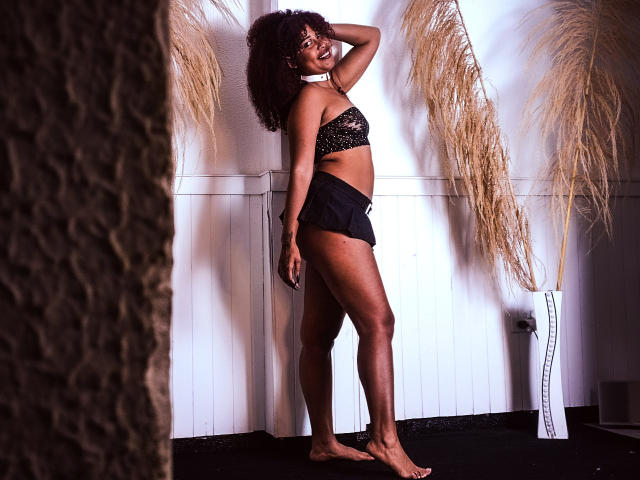 DalilaHeinz with curly hair, wearing a black sparkly bra and black shorts, standing barefoot, hand in hair, looking confident, tall and toned, with a sexy, curvy ass.