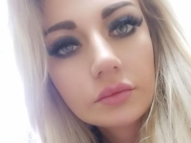 PenelopeAmore has blonde hair, blue eyes, thick black eyebrows, long black eyelashes, and pink lips, looking serious in a selfie. Her nose is small and she has a fair skin tone.