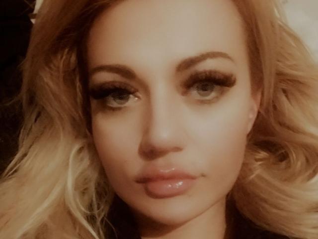 PenelopeAmore has blonde hair, full eyebrows, long eyelashes, and shiny pink lips. Her green eyes look slightly to the side with a subtle, confident expression.