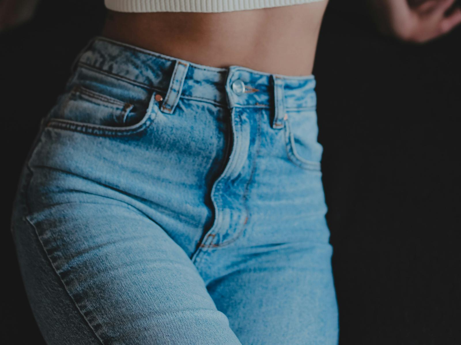 ImSweety's tight, blue, high-waisted jeans, showing their ass, with visible buttons and a white ribbed crop top, against a dark background.