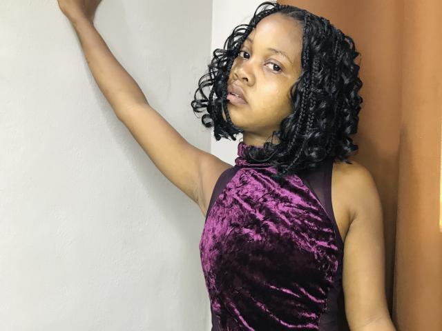 DareMagik with curly black hair, wearing a purple velvet halter dress, arm raised against a wall, looking slightly to the side with a neutral expression.