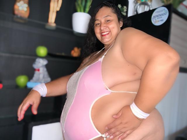 TistMature, una mujer latina de talla grande con cabello oscuro y rizado, vestida con un traje de baño entero rosa y blanco, que muestra senos grandes y un trasero prominente, sonriendo con lápiz labial rojo..