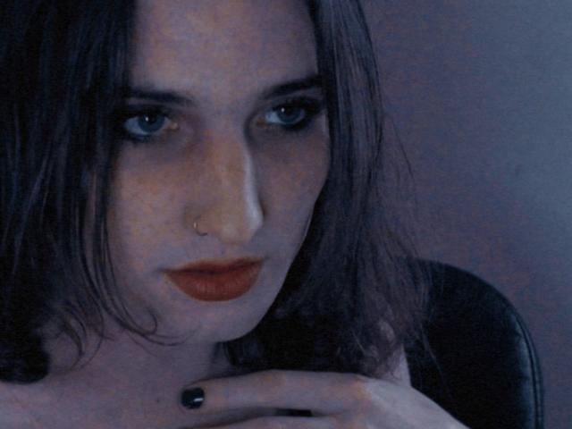 AlixeST has dark brown wavy hair, blue eyes, and a nose ring. She's wearing red lipstick and black nail polish, with a serious expression. Her hand is near her chest.
