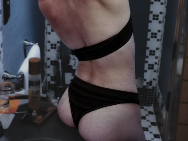 La espalda desnuda de AlixeST y su tanga negra, que muestran su trasero y sus glúteos pequeños y firmes, en un baño con una pared de azulejos. Su piel es pálida y lleva ropa interior negra..