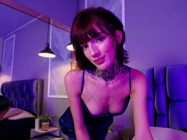 AmellieJones, with short brown hair and tattoos, leans forward in a blue dress, showing her tits and hairy chest, under purple lighting. Her face has a cute, subtle smile.