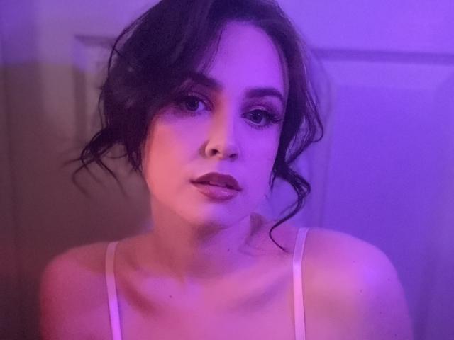 SultryRebel with dark wavy hair, purple-lit face, and pink lipstick, wearing a white spaghetti strap top, looking directly at the camera with a slight pout.
