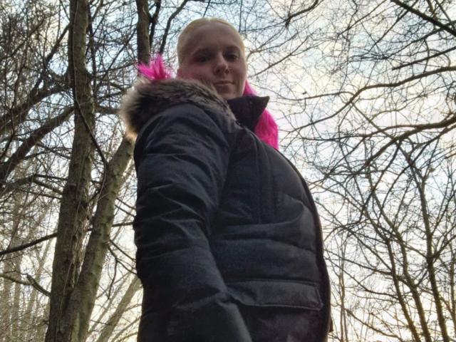Curvy-Sunny-hot in a black puffy jacket with furry hood, pink collar, and bald head, standing in a forest with leafless trees, looking down at the camera.