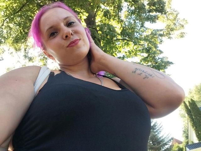 Curvy-Sunny-hot with pink hair, black tank top, and tattoo on right arm, holding head with left hand, standing outdoors, sunlight filtering through trees. Boobs visible, confident expression.