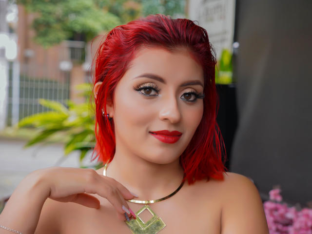 LizBonnet, avec ses cheveux roux éclatants, son rouge à lèvres rouge et son collier en or, sourit à l'appareil photo, sa main touchant sa poitrine, montrant ses seins, dans un décor urbain coloré..